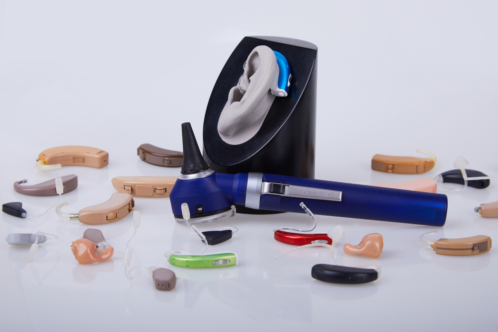 A blue otoscope and various hearing aids in different colors and styles are displayed around a model of an ear mounted on a stand, all set against a white background.