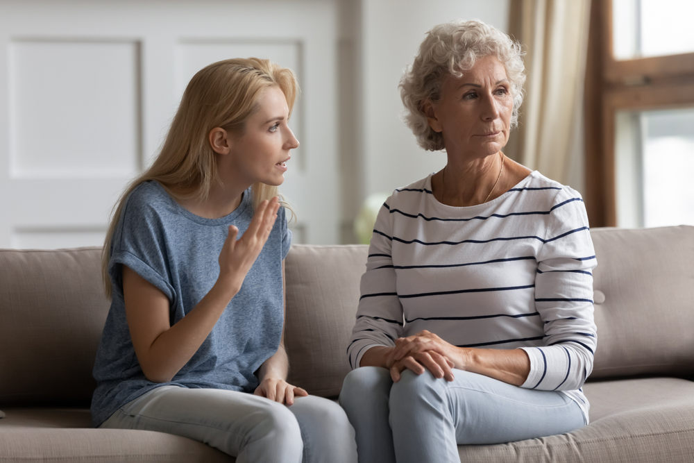 A younger woman is talking with concern to an older woman who looks upset and is looking away. They are sitting on a couch in a bright living room.