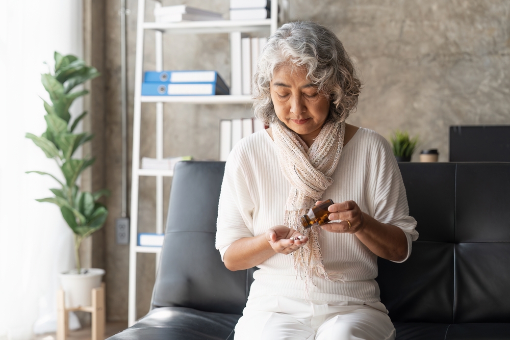 An older woman with gray hair sits on a black sofa, wearing a white top and scarf, looking down as she pours pills from a brown medicine bottle into her hand. A bookcase and plant are visible in the background.