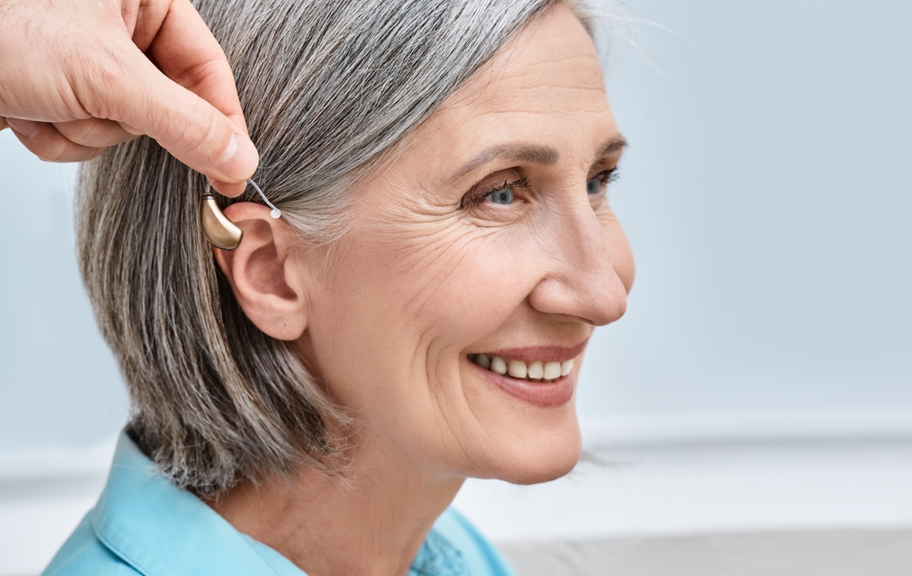 A smiling older woman with gray hair is having a behind-the-ear hearing aid placed by another persons hand.