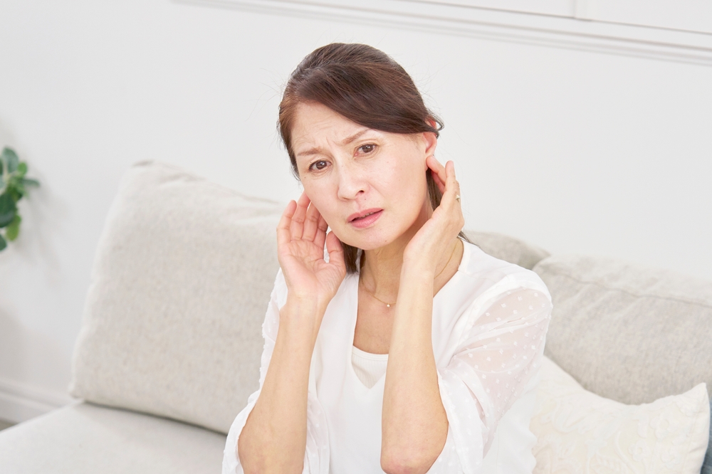 A woman sitting on a light-colored sofa touches both her ears with a concerned expression, suggesting she may be experiencing ear discomfort or pain.