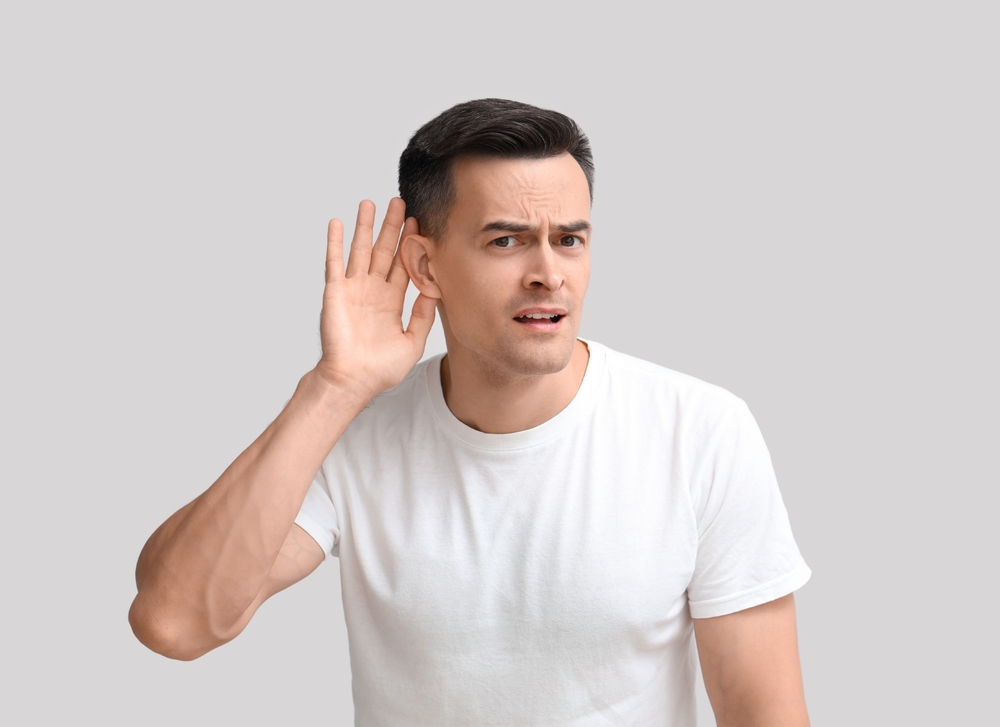 A man in a white t-shirt holds his hand to his ear and tilts his head, appearing to listen carefully or indicating he cannot hear well, against a plain light gray background.