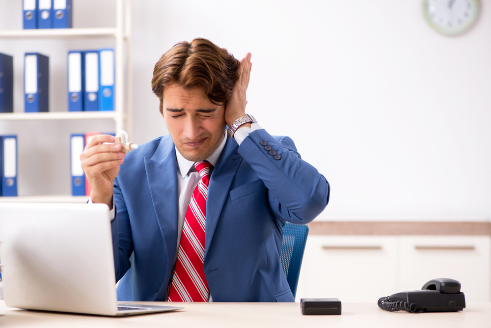 A man in a suit sits at an office desk holding a cup and covering one ear, appearing uncomfortable, with a laptop, phone, and office supplies on the desk.