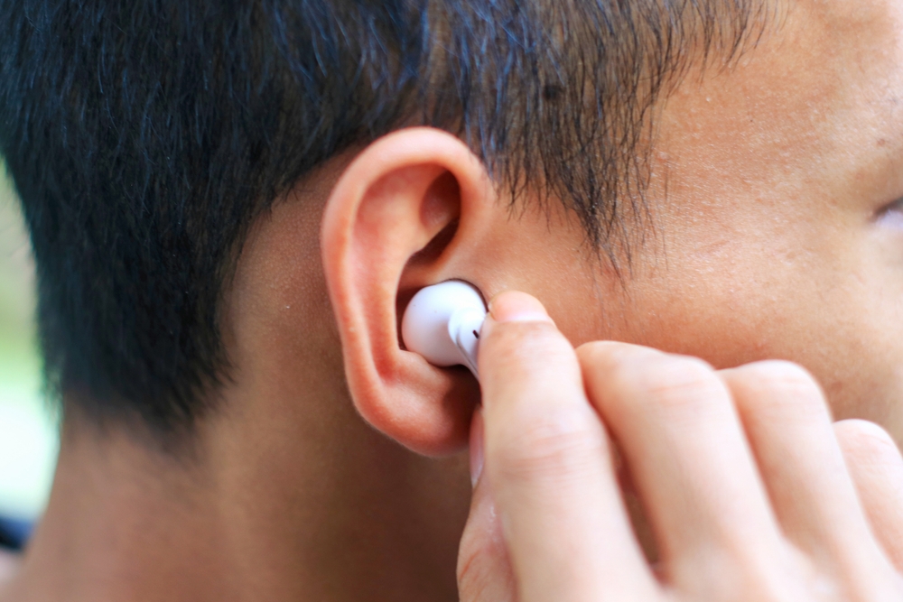 A person inserts a white wireless earbud into their ear, with their hand holding the earbud close to the ear. The background is blurred.