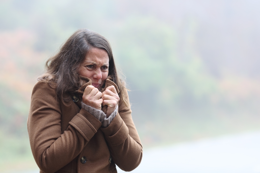 A woman wearing a brown coat stands outdoors, shivering and clutching her collar for warmth, with a misty, blurred background of trees.