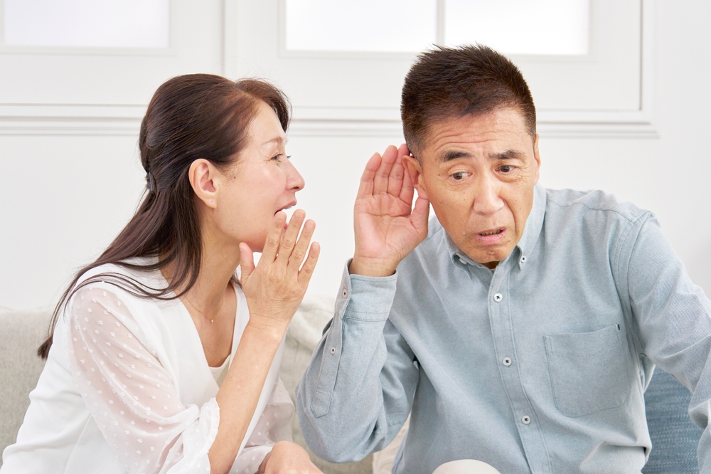 A woman in a white blouse whispers to a man in a light blue shirt, who is cupping his ear and leaning in to hear her better. They are sitting indoors on a light-colored sofa.